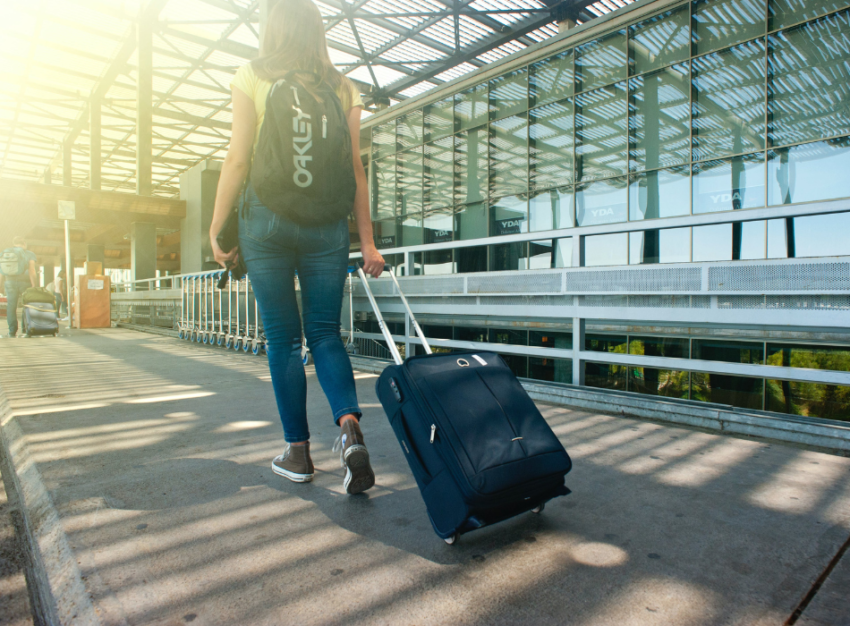 A woman walks down a sidewalk outside an airport. She is wearing jeans, a yellow t-shirt and a black backpack and pulling a navy blue wheeled suitcase.