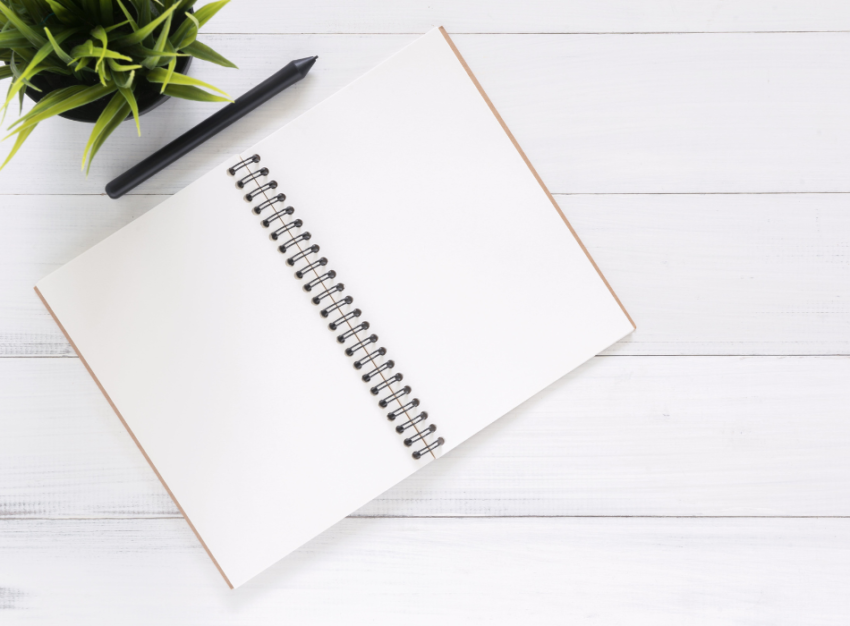 A blank white coil notebook sits open on a white surface with a black pen and plant in the top left corner