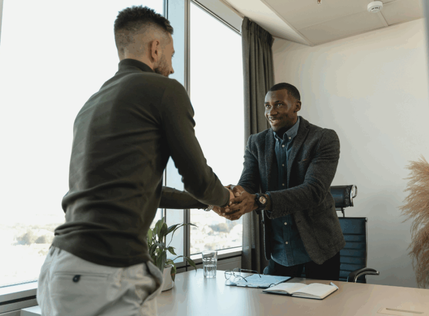 Two men shake hands across a desk.