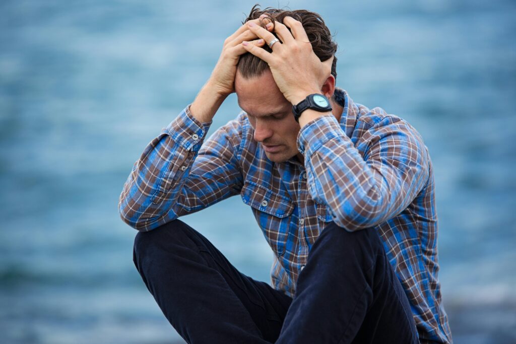 A stressed man in a blue plaid shirt sits and runs his hands through his hair.