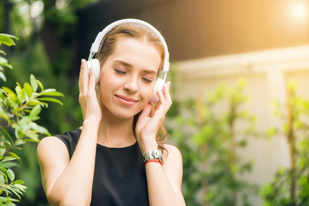 A girl listens to music from over-ear headphones in front of small trees.