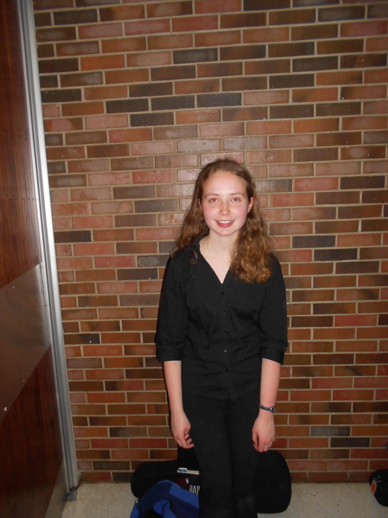 A girl dressed all in black stands in front of a brick wall, a violin case at her feet.
