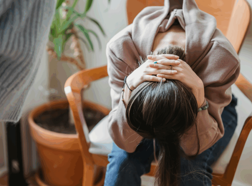 A woman slumps in a chair with her head bent over and her hands clasped behind her hair.