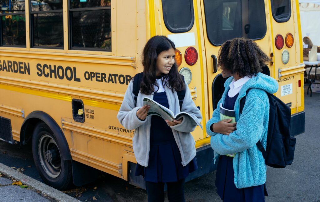 Two girls wearing backpacks stand behind a yellow school bus.
