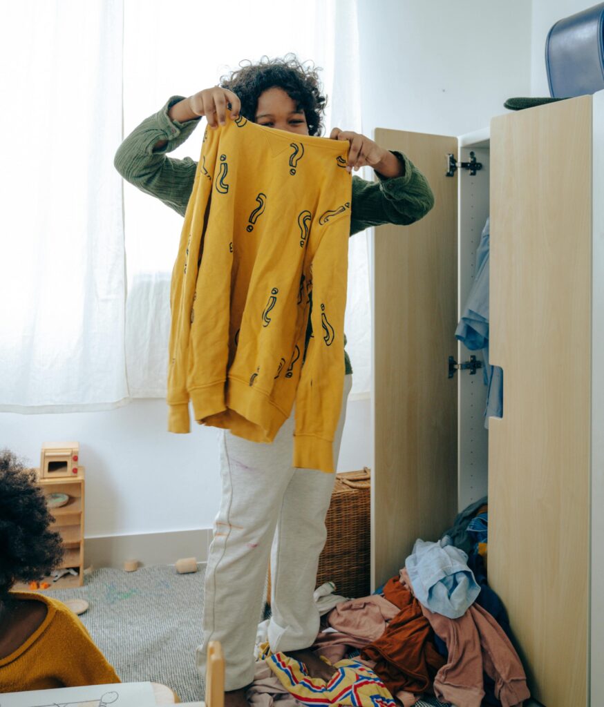 A boy holds up a yellow shirt with question marks on it above a pile of clothes on the floor.