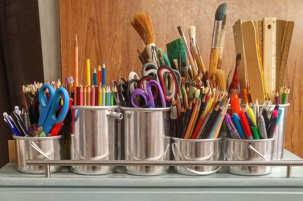 A set of small silver buckets containing rulers, paint brushes, coloured pencils, scissors and other school supplies.