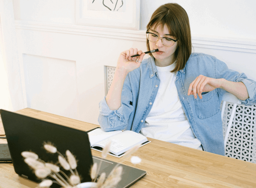 A girl wearing a jean jacket holds a pen while staring down at a notebook with a laptop in front of her.