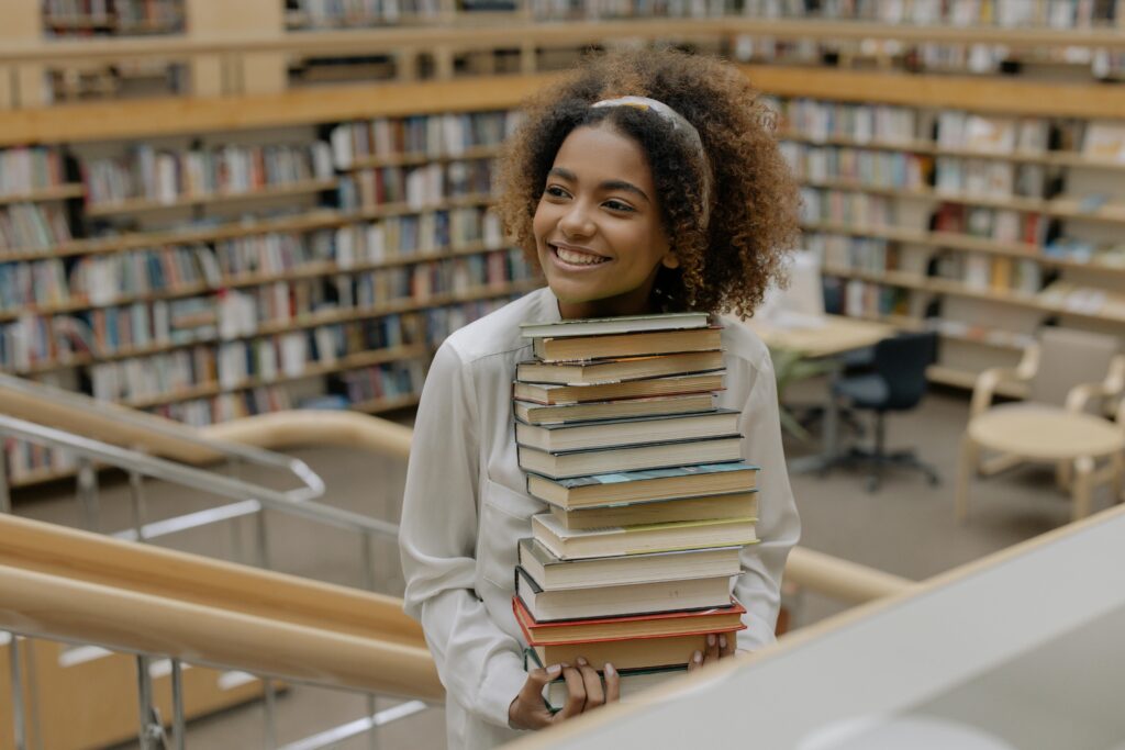 A girl holds a tall stack of books with the interior of a library in the background.