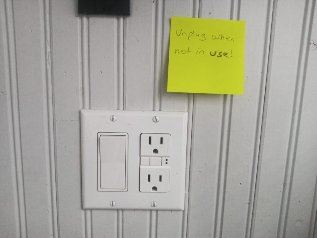 A wall outlet with a sticky note reminder to unplug items when not in use.