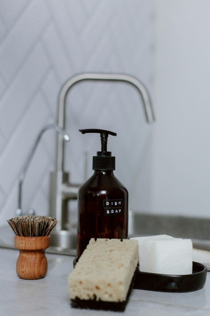A dish sponge, white block, bottle of dish soap and wooden dish brush sit next to a sink.