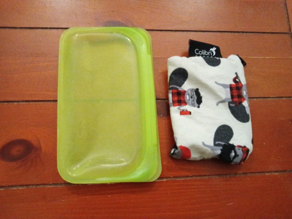 A green silicone snack bag and a fabric snack bag featuring beavers in red plaid shirts sit on a wooden surface.
