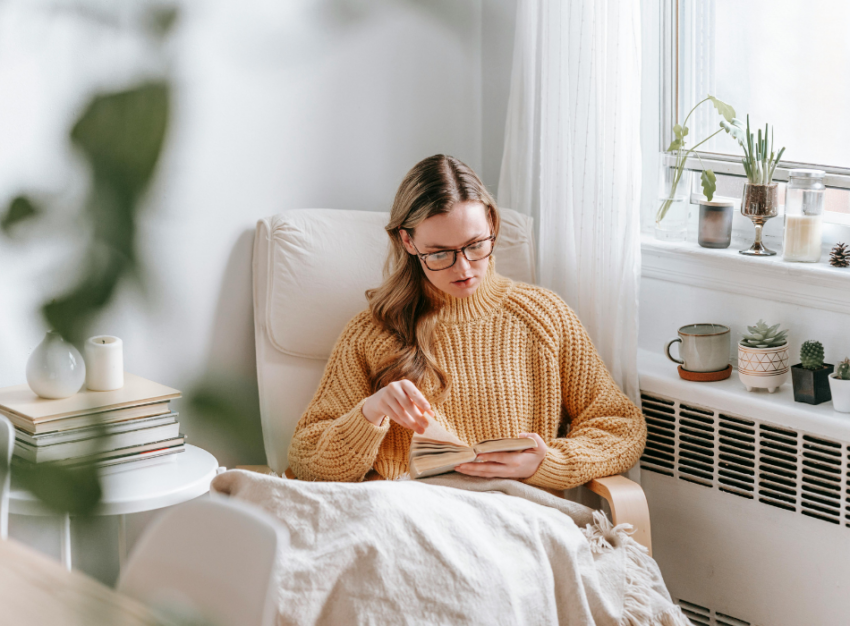 A woman in a pale orange sweater sits reading a book wrapped up in a blanket.