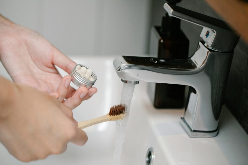A person wets a bamboo toothbrush under a running tap while holding a small metal tin containing toothpaste tablets.